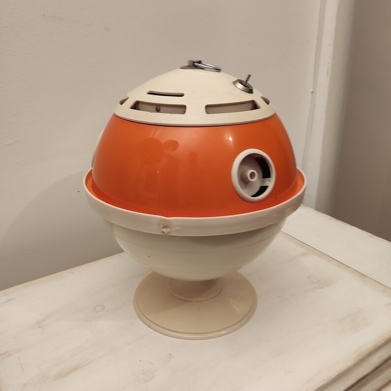May include: Orange and white egg-shaped vintage ashtray with a metal ring on top. The ashtray has a round base and a small hole on the side.