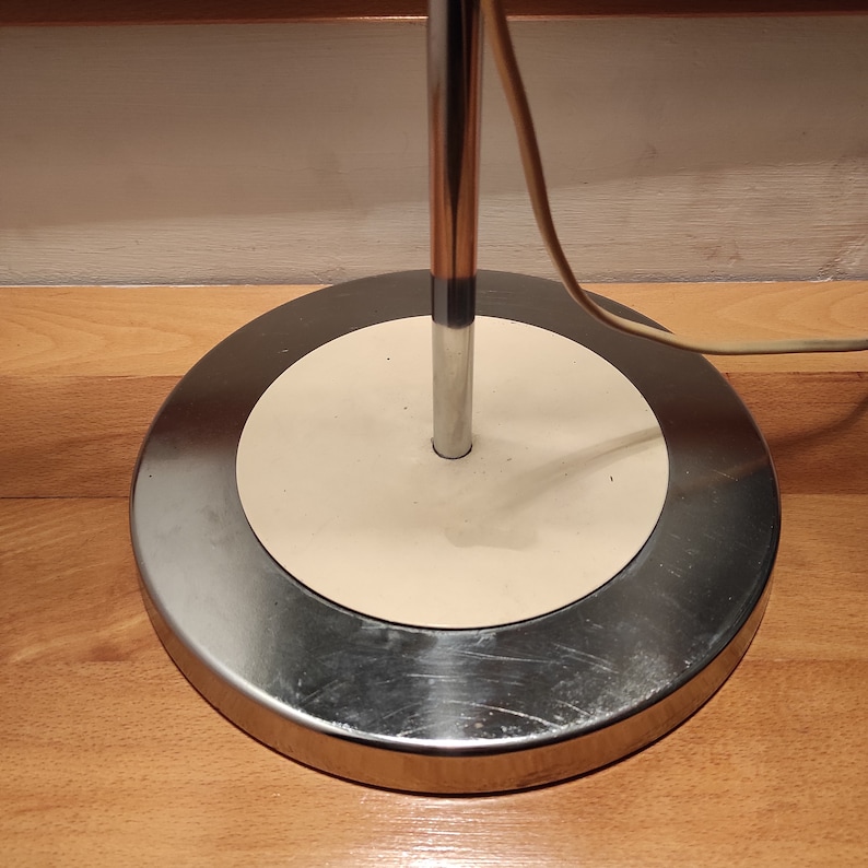 May include: A chrome floor lamp with a white circular base. The lamp has a single light bulb and a cord that plugs into an outlet.
