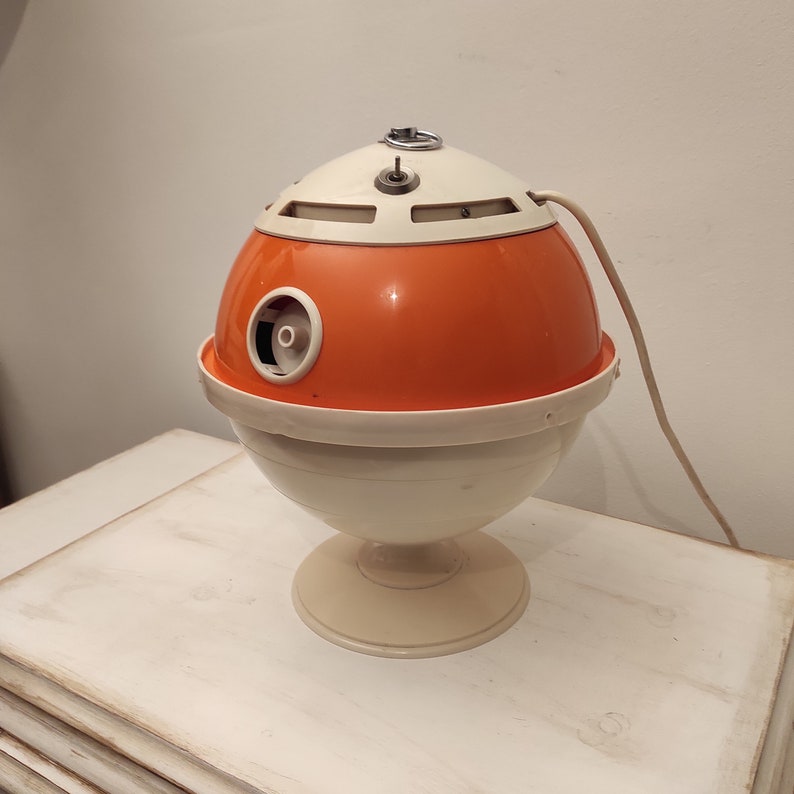 May include: A vintage orange and white electric hair dryer with a round, futuristic design. The dryer has a cord and a base.