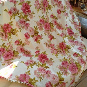 Puede incluir: Un mantel de tela blanco con un patrón floral rosa y verde. El mantel tiene un borde con volantes.