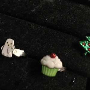 Könnte beinhalten: Eine Sammlung von ausgefallenen Ringen mit skurrilen Designs. Enthält einen weißen Geisterring, einen Cupcake-Ring mit grünem Boden und roter Kirsche sowie einen grünen Weihnachtsbaumring mit roten Akzenten. Vor schwarzem Hintergrund.