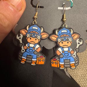 Puede incluir: Un par de pendientes novedosos con personajes de vacas de dibujos animados vestidos de mecánicos. Cada vaca lleva un gorro azul y un mono, sosteniendo una llave inglesa y una caja de herramientas. Los pendientes tienen ganchos dorados.