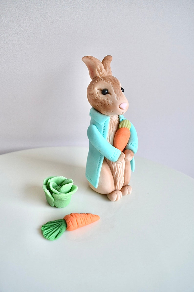 May include: A blue-coated fondant bunny rabbit holding a carrot, with a green fondant cabbage and carrot on a white surface.