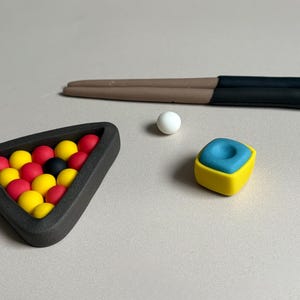 Könnte beinhalten: Ein Miniatur-Billard-Set mit einem Dreieck aus roten, gelben und schwarzen Billardkugeln. Eine weiße Kugel, ein blau-gelber Kreidewürfel und ein braun-schwarzer Billardqueue sind ebenfalls vorhanden.