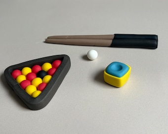May include: A miniature pool table set with a triangle rack of red, yellow, and black pool balls. A white cue ball, a blue and yellow chalk cube, and a brown and black pool cue are also present.