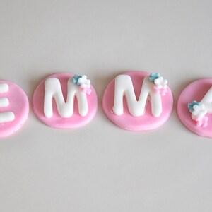 May include: Pink fondant cake toppers with white letters spelling out the name "EMMA". Each letter has small blue and pink flowers on top.