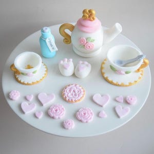 May include: A decorative tea set featuring a white teapot, teacups, and saucers, adorned with pink roses and gold trim. The set includes a small blue bottle with a tag reading "Drink Me". Additional pink hearts and flowers are scattered around.