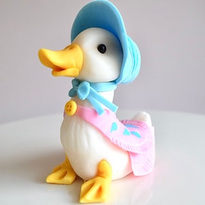 May include: A white duck figurine wearing a blue bonnet and a pink dress with blue accents. The duck has yellow feet and a yellow beak.
