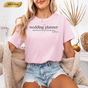 Può includere: Una maglietta rosa chiaro con la scritta "wedding planner, professional problem solver" in nero. La maglietta è a maniche corte e ha uno scollo tondo. Le parole "Soft Pink" sono in un riquadro dorato in alto a sinistra.