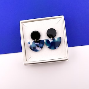 Op de afbeelding: Een paar oorbellen met een zwarte cirkelvormige bovenkant en een halfronde onderkant. De onderste helft heeft een blauw-wit marmerpatroon. De oorbellen worden gepresenteerd in een witte doos tegen een blauwe achtergrond.