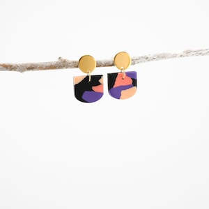 Könnte beinhalten: Ein Paar goldene und schwarze Ohrringe mit geometrischem, abstraktem Design. Die Ohrringe haben ein schwarzes, violettes und pfirsichfarbenes Farbmuster.