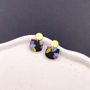 Puede incluir: Un par de pendientes negros semicirculares con patrones abstractos coloridos en morado, amarillo y azul. Cada pendiente tiene un pequeño perno circular dorado en la parte superior. Los pendientes se exhiben sobre una superficie blanca y curvada.