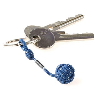 Puede incluir: Un llavero de cuerda azul con una bola anudada en el extremo. El llavero está unido a dos llaves plateadas.