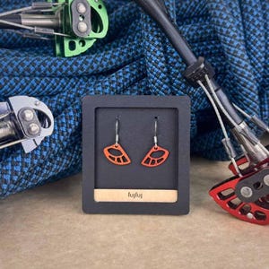 Könnte beinhalten: Offene, orangefarbene Ohrringe mit silbernen Haken, präsentiert in einer schwarzen Box. Die Ohrringe haben eine einzigartige, geometrische Form. Im Hintergrund befinden sich Kletterausrüstung und ein blaues Seil.