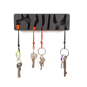 Puede incluir: Un porta llaves negro con cinco ganchos, cada uno con una llave diferente. El porta llaves está hecho de un material texturizado y tiene un diseño moderno. Las llaves son plateadas y tienen diferentes formas y tamaños.