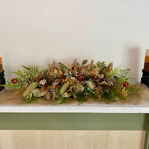 May include: A fall-themed mantel arrangement featuring a central floral display with faux berries, leaves, and greenery on a burlap runner. Two pillar candles in shades of brown and orange sit on black candle holders on either side.