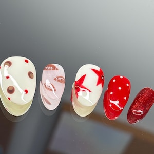 Könnte beinhalten: Ein Set aus fünf künstlichen Nägeln mit verschiedenen Designs. Die Nägel zeigen eine cremefarbene Basis mit braunen Flecken und roten Punkten, einen rosa Nagel mit einem Football-Design, einen weißen Nagel mit einem roten Stern, einen roten Nagel mit weißen Punkten und einen glitzernden roten Nagel.