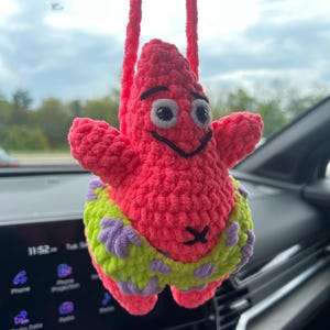 May include: A crocheted hanging ornament shaped like a pink starfish character with black embroidered eyes and a mouth. The figure wears lime green shorts featuring a purple floral pattern and is suspended by a matching pink crocheted yarn loop.