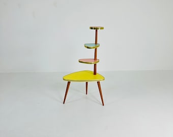 Könnte beinhalten: Ein gelber, rosa und blauer Pflanzenständer im Mid-Century-Stil mit drei Ablagen. Der Ständer hat eine dreieckige Basis und Holzbeine.