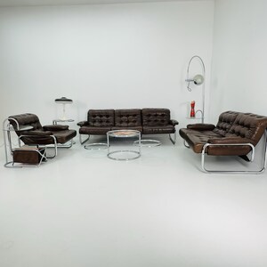 May include: A brown leather sofa with chrome legs and arms, two matching armchairs, and three chrome and glass coffee tables. The sofa and armchairs have tufted upholstery.