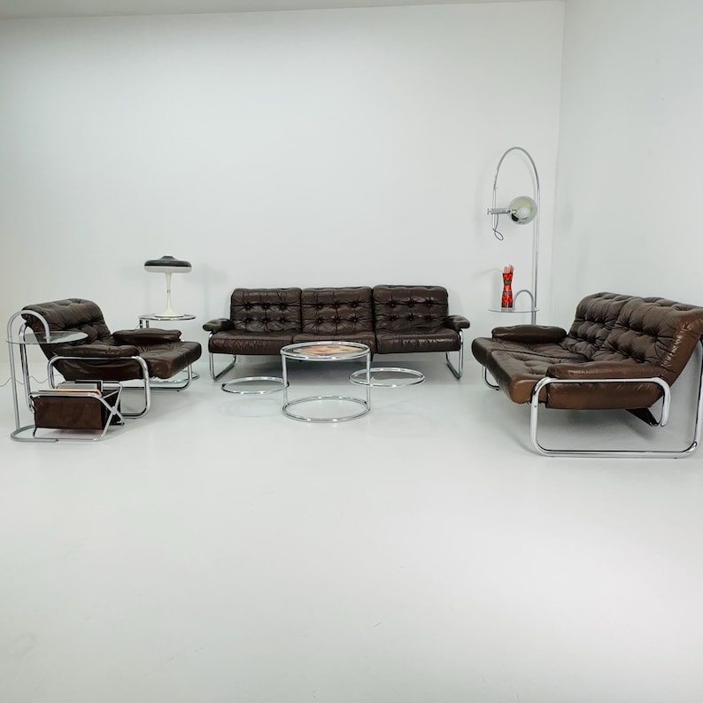 May include: A brown leather sofa with chrome legs and arms, two matching armchairs, and three chrome and glass coffee tables. The sofa and armchairs have tufted upholstery.