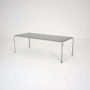 Peut inclure: Une table basse rectangulaire dotée d'un plateau en verre foncé semi-transparent, soutenu par une structure en métal chromé poli. L'ensemble se compose de quatre pieds tubulaires reliés par un cadre métallique continu supportant le verre.