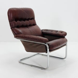 May include: A vintage brown leather armchair with a chrome frame. The chair features a high back, padded armrests, and a tufted design on the seat and backrest. The chair is designed with a cantilevered base.
