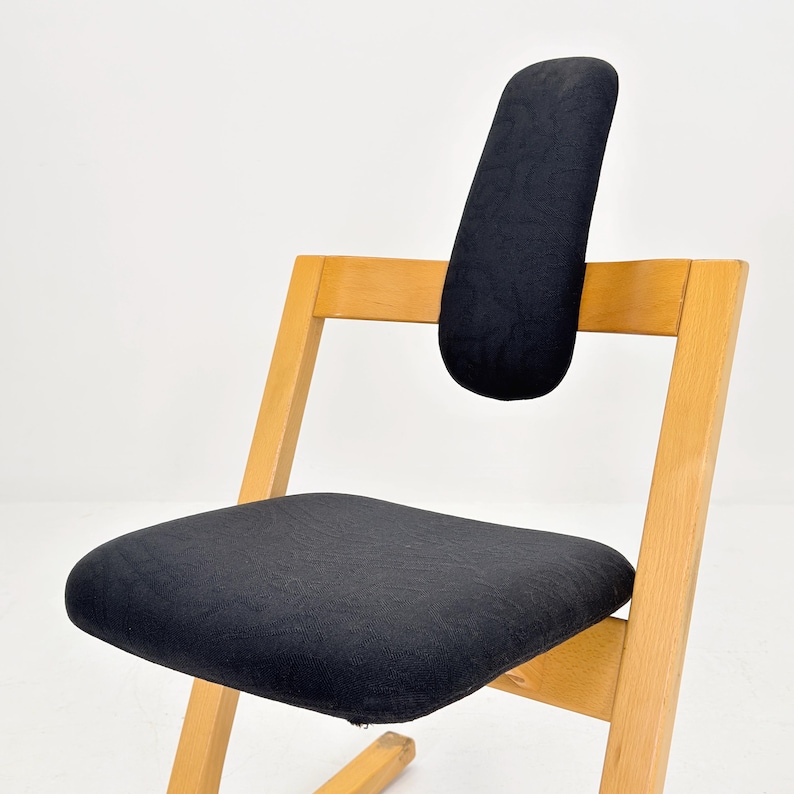 May include: A wooden chair with a black seat and backrest. The chair has a curved backrest and a slanted seat. The frame is made of light-colored wood. The chair is designed for comfort and support.