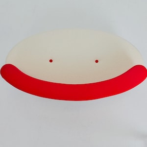 K&ouml;nnte beinhalten: Eine ovale, cremewei&szlig;e Schale mit strukturierter Oberfl&auml;che und einem glatten, leuchtend roten Rand entlang der Vorderseite. Im Inneren der Schale befinden sich zwei kleine, deutlich rote, kreisf&ouml;rmige Akzente, die symmetrisch zur Mitte hin angeordnet sind.