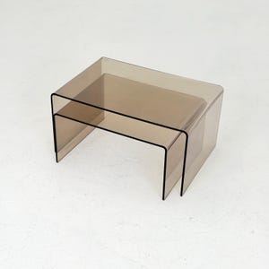 以下が含まれることがあります： 半透明のスモークカラーのアクリル製ネストテーブル2点セット。各テーブルは、滑らかで丸みを帯びたエッジを持つ連続したU字型のカーブデザインが特徴で、小さいテーブルを大きいテーブルの下にきれいに収めることができる構造になっています。