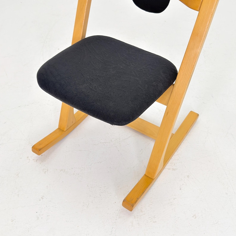 May include: A wooden chair with a black seat and backrest. The chair has a light-colored wooden frame and a simple, minimalist design. The seat and backrest are made of a dark, textured fabric. The chair is angled slightly, suggesting a comfortable seating position.