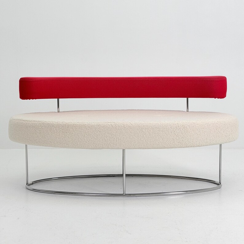 K&ouml;nnte beinhalten: Ein kreisf&ouml;rmiges Sofa mit einer cremefarbenen, strukturierten Sitzfl&auml;che und einer d&uuml;nnen, horizontalen roten R&uuml;ckenlehne. Das M&ouml;belst&uuml;ck wird von einem schlanken, ovalen Chrommetallrahmen getragen, der die Basis mit der R&uuml;ckenlehnenst&uuml;tze verbindet.