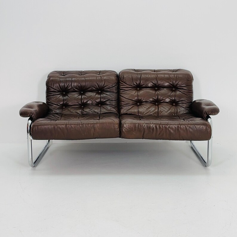 May include: A brown leather sofa with a tufted design and chrome legs. The sofa has a modern style and is perfect for a living room or den.