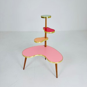 Può includere: Un portavasi a tre livelli di colore rosa con finiture dorate. Il portavasi ha una forma unica e curva ed è realizzato in legno con una finitura marrone chiaro.