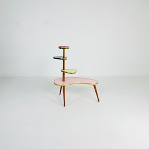 May include: A mid-century modern plant stand with four tiers. The stand is made of wood with a pink, cream, and blue finish. The stand has a unique, organic shape and is perfect for displaying plants or other decorative items.