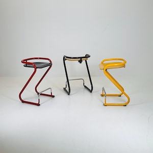 May include: Three Z-shaped bar stools with black, red, and yellow metal frames. The stools have a simple design with a curved backrest and a footrest.