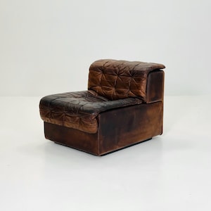 May include: A vintage, low-profile armchair in distressed brown leather. The seat and backrest feature a quilted pattern, and the armrest is integrated into the design. The chair has a boxy shape and a dark brown base, showcasing a mid-century modern aesthetic.