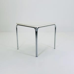 May include: A small, square side table with a white top and chrome legs. The table is approximately 50 cm wide and 50 cm deep, with a height of about 50 cm. The legs are curved and tubular, giving the table a modern look.