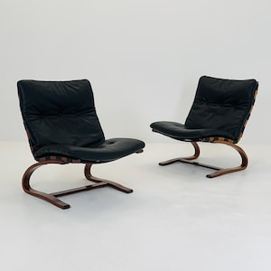 May include: Two mid-century modern lounge chairs. Each chair features a curved wooden frame, a black leather seat, and a black leather backrest. The backrests have woven wood accents. The chairs are set against a white background.