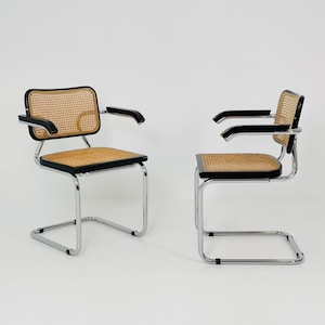 Puede incluir: Dos sillones cantilever vintage. Cada silla presenta respaldo y asiento de caña tejida, ribete negro y estructura de acero cromado. Los reposabrazos son negros. Las sillas están sobre un fondo blanco.