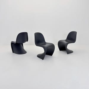 May include: Three black, sculptural chairs with a distinctive, flowing, S-shaped design. The chairs are made of a solid, matte material and are set against a plain white background. The chairs have a modern, minimalist aesthetic.
