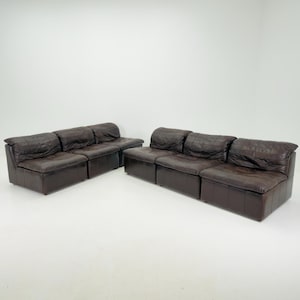 Könnte beinhalten: Ein modulares Sofaset bestehend aus zwei separaten Abschnitten mit jeweils drei verbundenen Sitzen. Die Möbel sind mit dunkelbraunem Leder bezogen, das sichtbare Nähte und eine weich gepolsterte Optik an Rücken- und Sitzflächen aufweist.