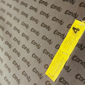 May include: A close-up of a brown fabric with the word "Candy" repeated in a pattern. A yellow tag with the text "Warenpass bitte nicht entfernen" and "5191516665701" is attached to the fabric.