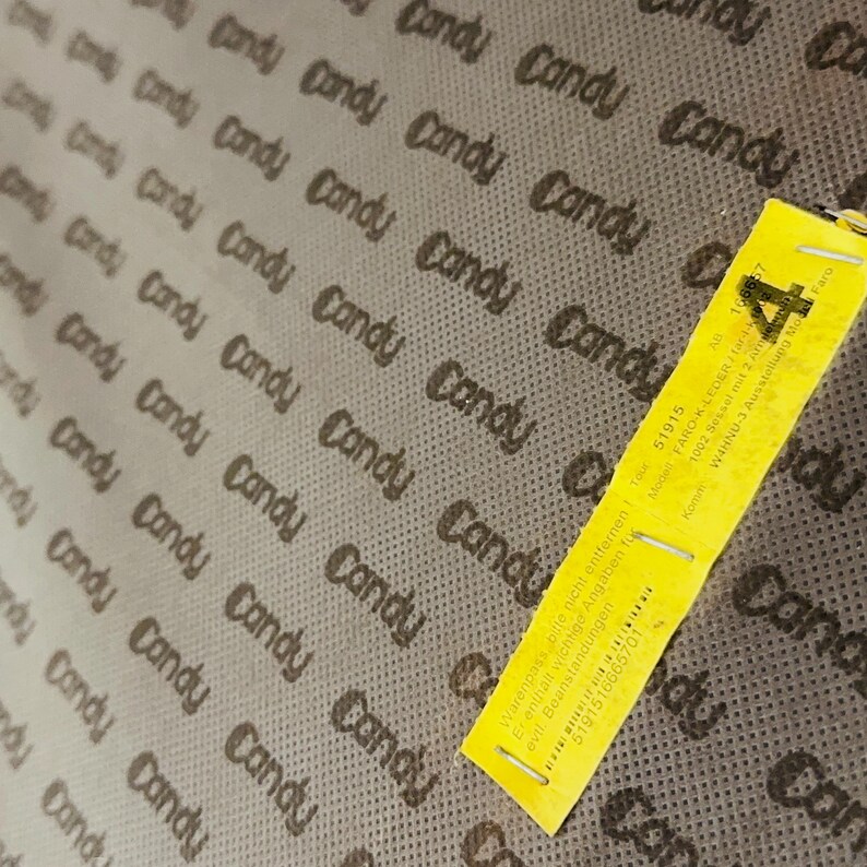 May include: A close-up of a brown fabric with the word "Candy" repeated in a pattern. A yellow tag with the text "Warenpass bitte nicht entfernen" and "5191516665701" is attached to the fabric.