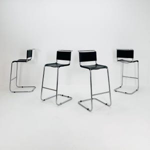 Puede incluir: Cuatro taburetes de bar negros y cromados sobre un fondo blanco. Los taburetes tienen un diseño minimalista, con asientos y respaldos negros, y marcos de metal cromado. Adecuados para una cocina o bar.