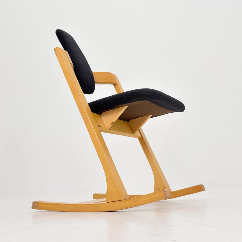 May include: A wooden rocking chair with a black seat and backrest. The chair has a unique, angled design with a curved base for rocking. The frame is made of light-colored wood, and the seat appears to be upholstered.