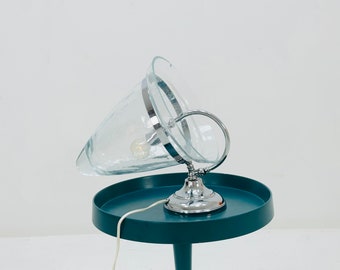 May include: A teal side table with a glass lamp with a chrome base. The lamp shade is clear glass and is tilted to the side.