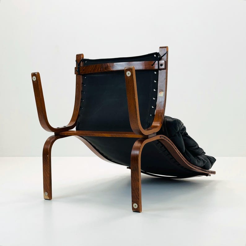Puede incluir: Un sill&oacute;n lounge de cuero negro con estructura de madera. El sill&oacute;n tiene un respaldo curvo y un asiento bajo. El sill&oacute;n est&aacute; hecho de madera oscura y tiene un coj&iacute;n de cuero negro.