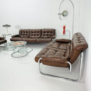 May include: A brown leather sofa with chrome legs and a matching armchair. The sofa has a tufted design and the armchair has a curved back. There are two glass and chrome coffee tables in front of the sofa.