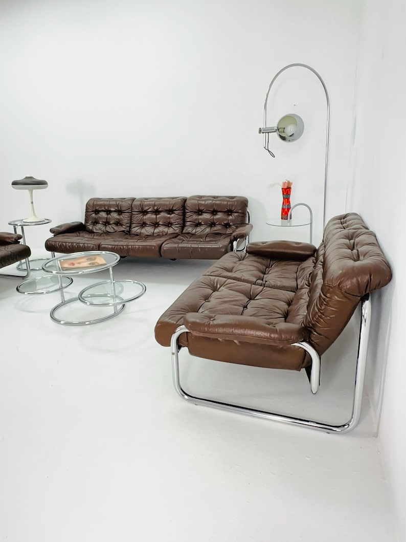May include: A brown leather sofa with chrome legs and a matching armchair. The sofa has a tufted design and the armchair has a curved back. There are two glass and chrome coffee tables in front of the sofa.